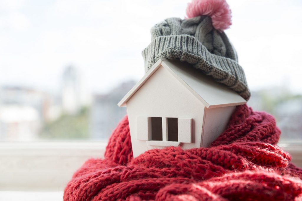 Home insulation may be the primary factor in keeping your home warm during winter. A toy wood house wrapped in a knit hat and scarf.