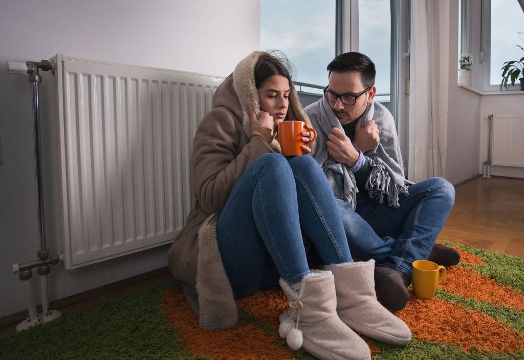 A freezing cold couple sits on the floor by their radiator. If you're too cold in your house, your furnace not turning on may be the issue.